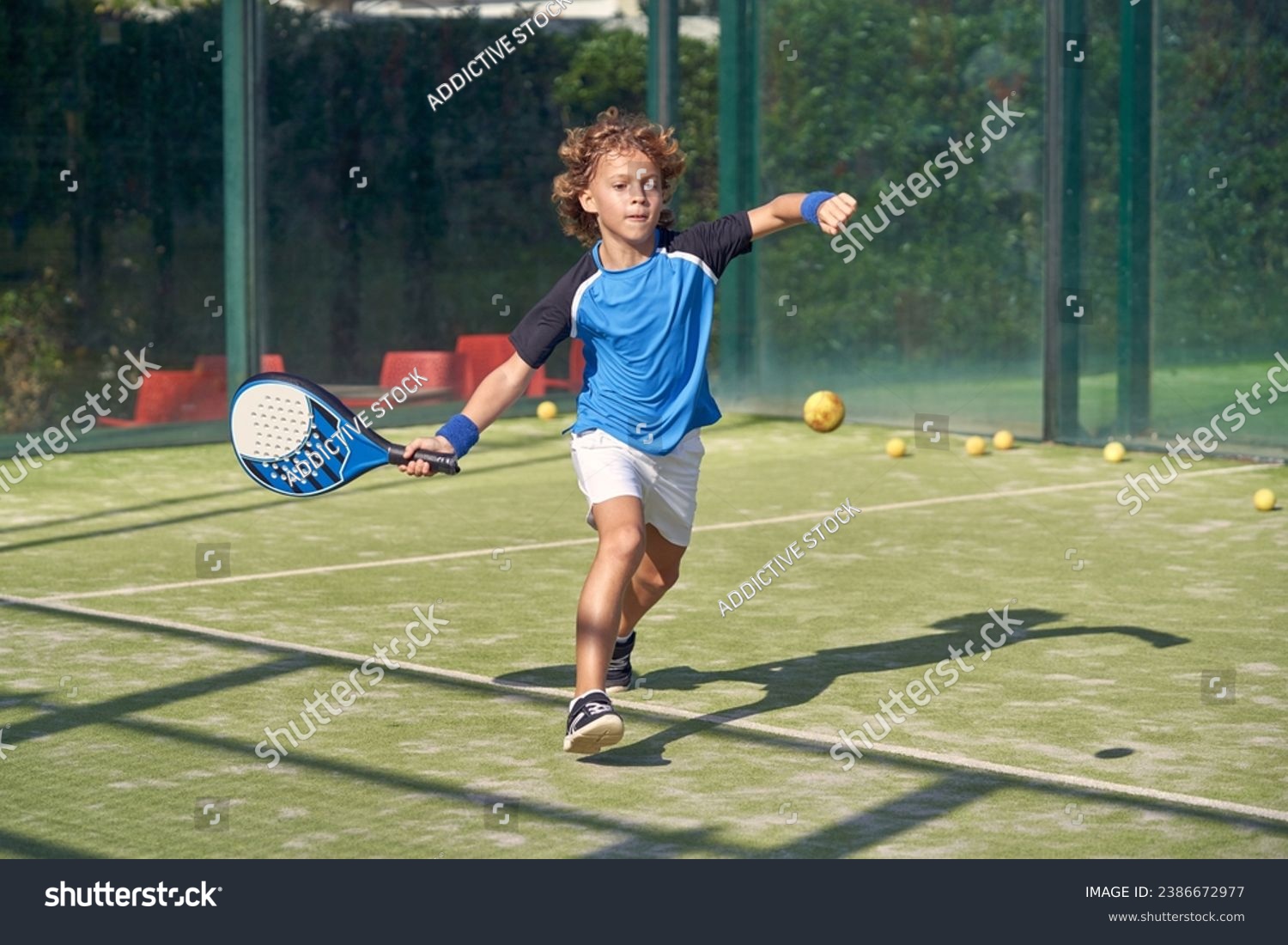 Full body of active boy in activewear hitting ball with racket while playing padel game on sports ground on summer day #2386672977