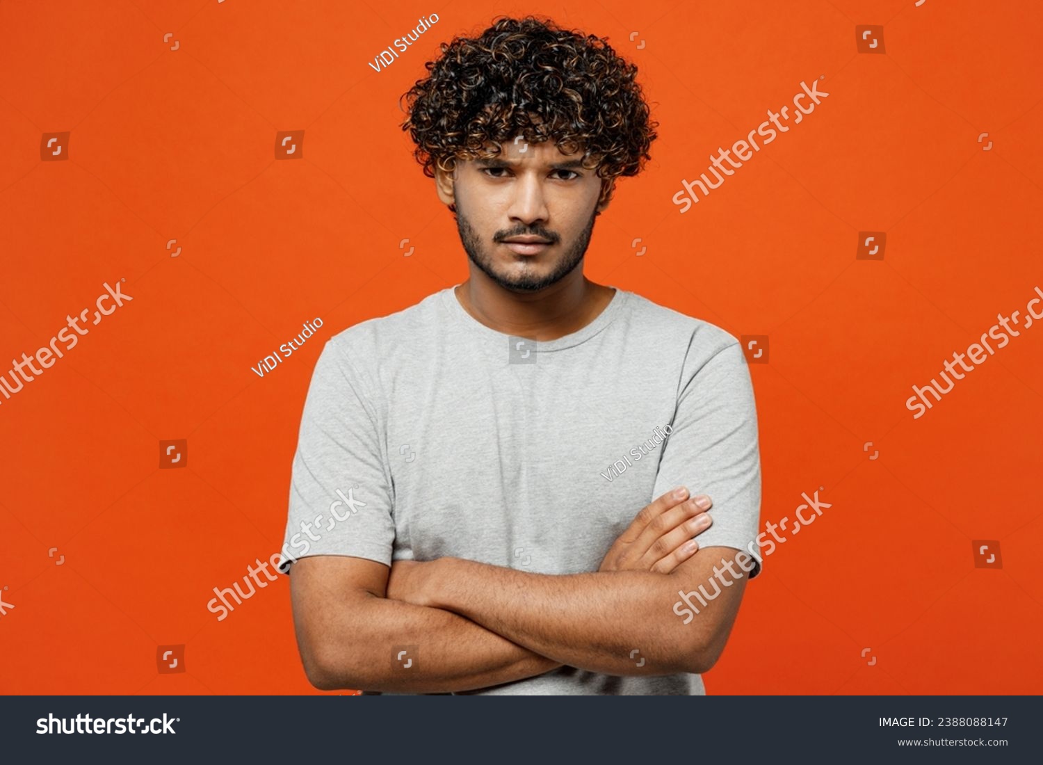 Young sad frowning mad furious shrewd Indian man he wearing t-shirt casual clothes hold hands crossed folded looking camera isolated on orange red color background studio portrait. Lifestyle concept #2388088147