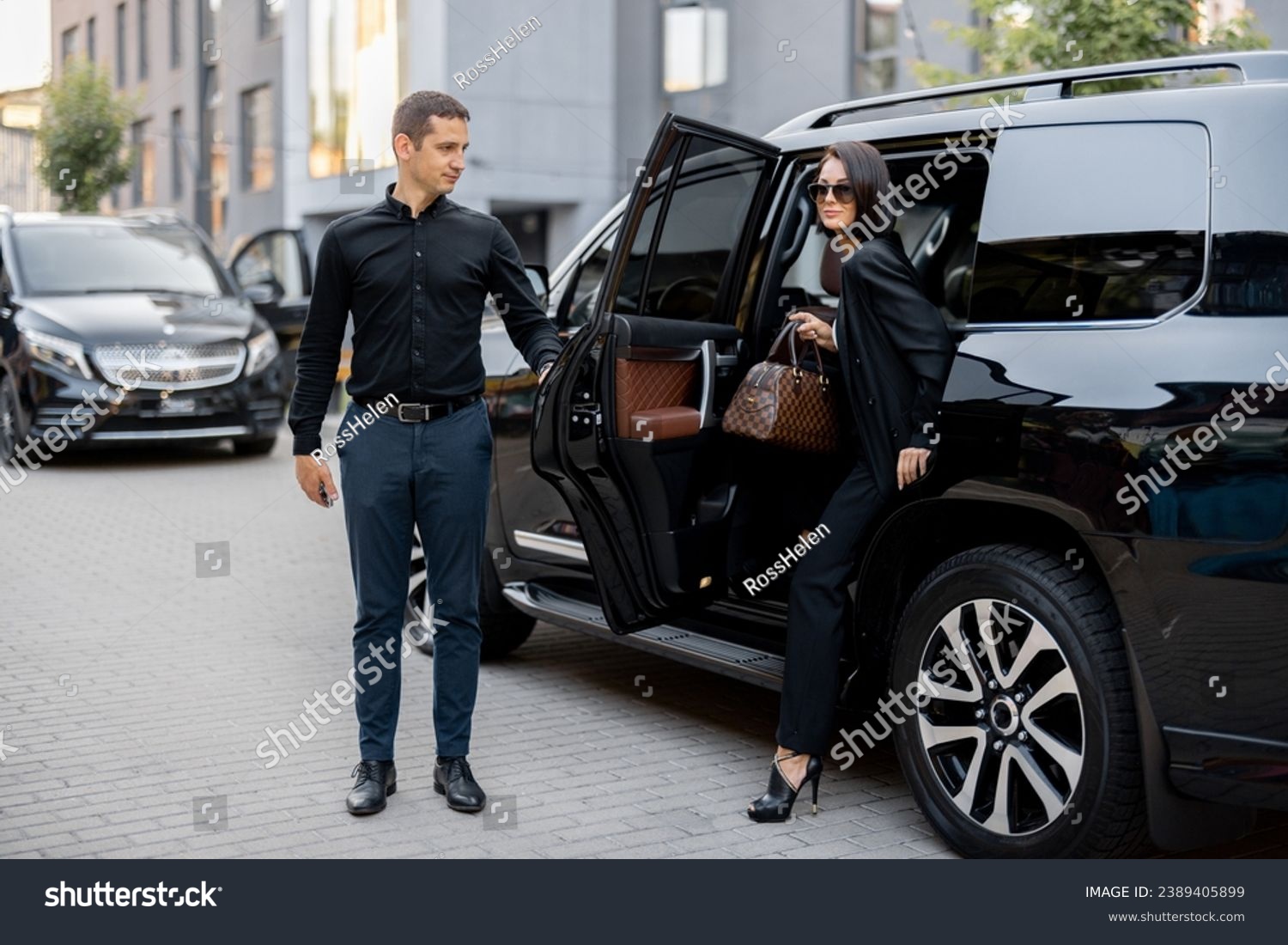 Male driver helps a business lady to get out of a car, opening door of a luxury SUV taxi. Business lady with handbag wearing black formal wear. Concept of transportation service #2389405899