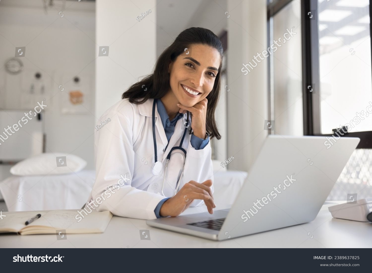 Cheerful attractive doctor woman professional portrait. Young Latin practitioner sitting at workplace table with laptop, looking at camera with toothy smile posing for shooting in surgery office #2389637825