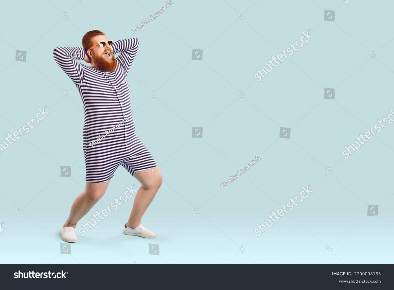 Funny fat man in swimwear having fun on the beach during summer holiday. Happy cheerful excited plump young guy wearing striped swimsuit or underwear posing on light blue copy space studio background #2390098163