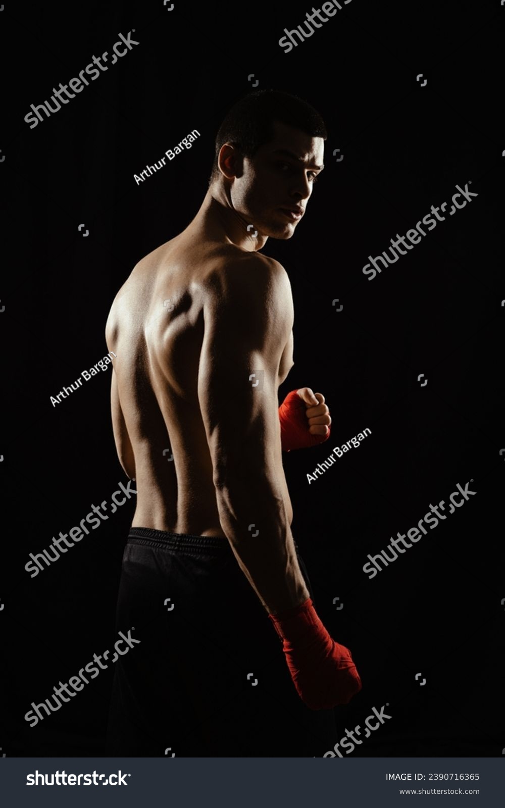 Portrait of male boxer posing in boxing stance, looking over shoulder, against black background. #2390716365