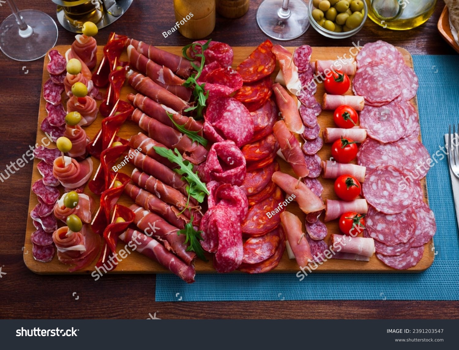 Top view of traditional Spanish meat platter - sliced dry-cured jamon, bacon and sausages on wooden board with olives, tomatoes and greens #2391203547