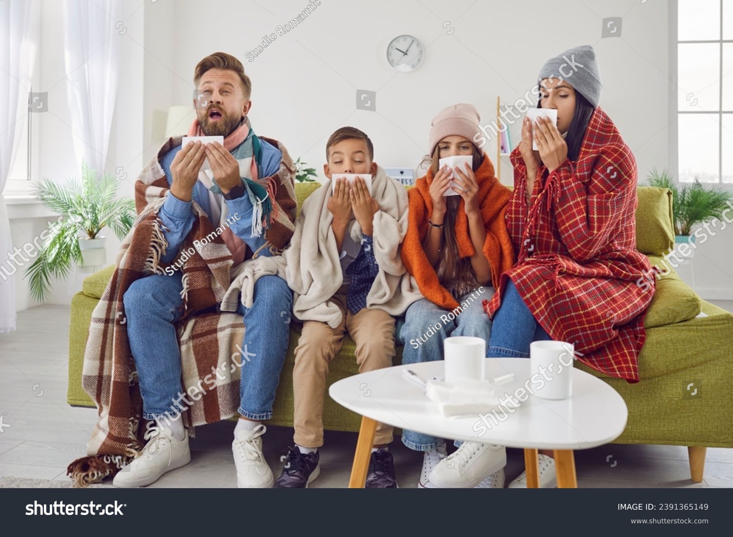 Achoo. Family of four have caught bad cold all together. Sick mom, dad and children in warm clothes and plaids sitting on couch in living room and sneezing in paper handkerchiefs. Winter, flu concept #2391365149