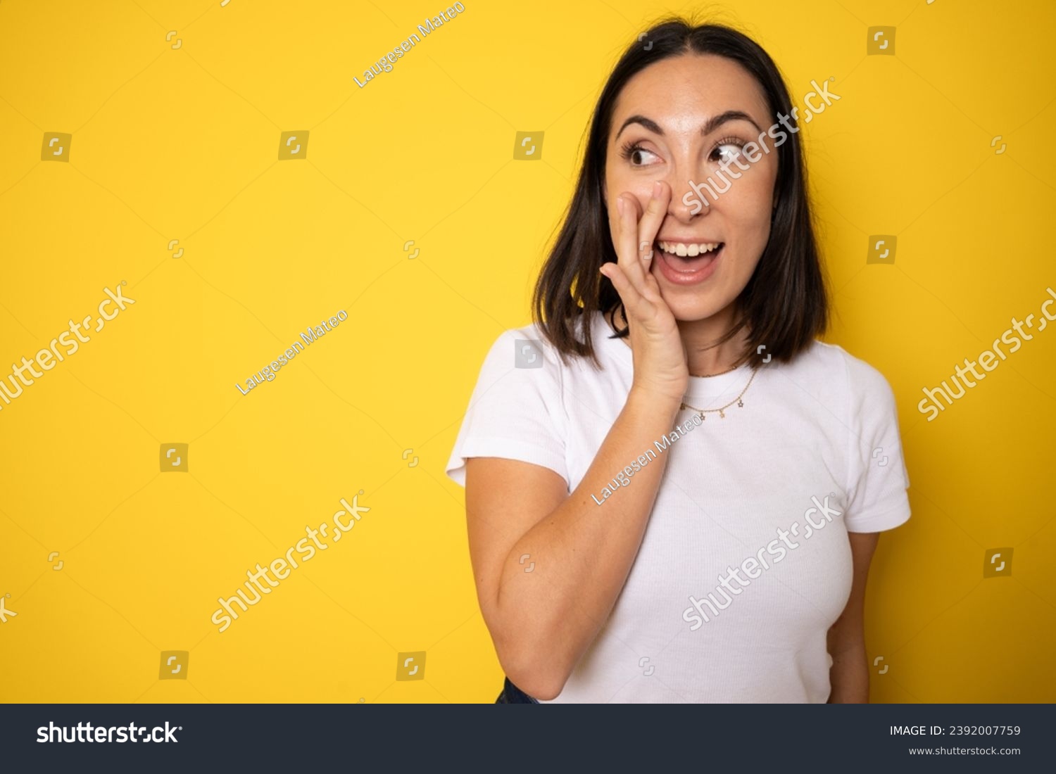 A close up portrait of a young pretty woman telling secret information, holding her hand near mouth and standing against yellow background #2392007759
