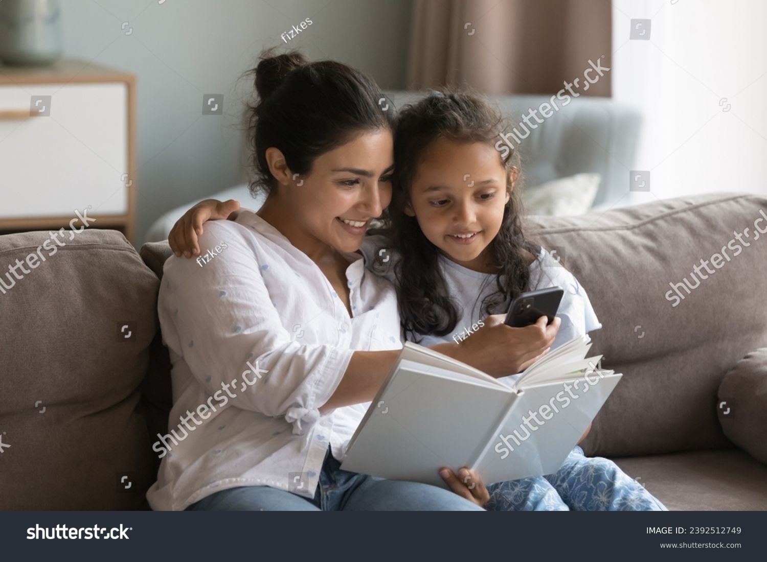 Indian mother and preschooler daughter sit on sofa at home holding storybook, distracted from reading use modern smartphone, search or buy books online via web store library, on-line bookshop concept #2392512749