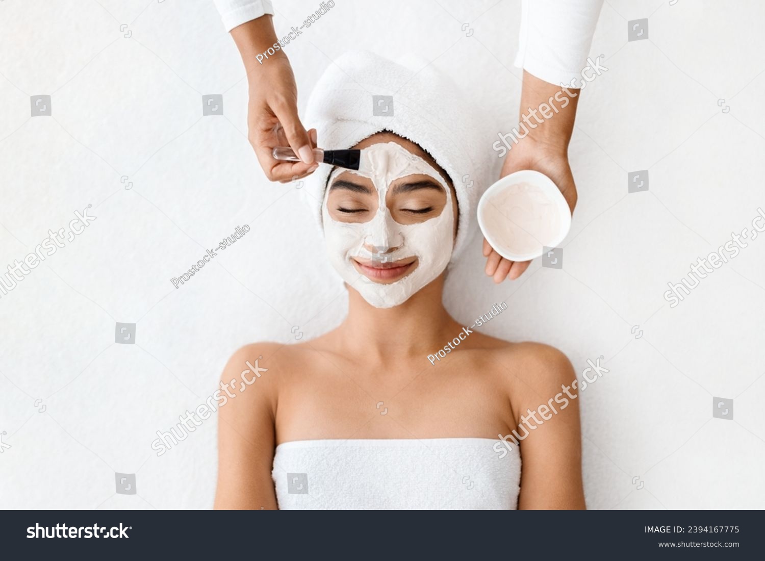 Top view of cosmetologist applying clay mask on face of young indian woman, beautiful eastern lady with towel on head lying on table, enjoying beauty treatments in spa salon, white background #2394167775