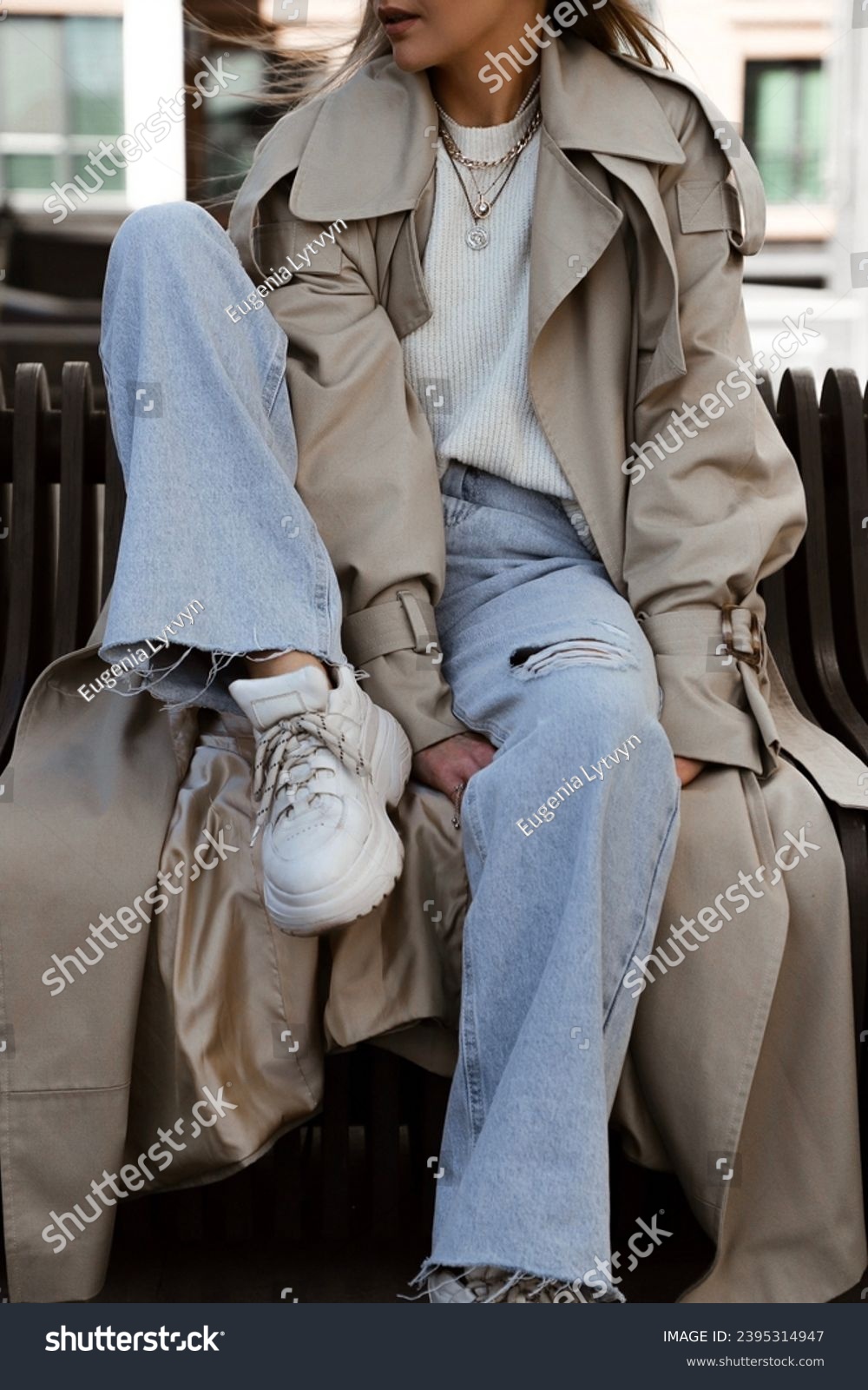 beautiful blonde girl dressed in beige oversized trench coat, blue palazzo jeans, white sweater, massive sneakers and bag, details of stylish fashion outfit, lifestyle model sitting #2395314947