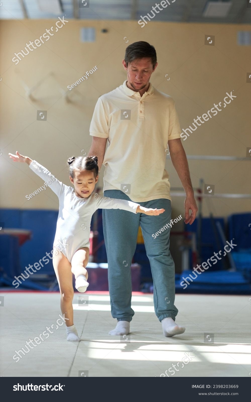 Full shot of cheerful Asian little girl wearing white bodysuit exercising in gymnastics class while Caucasian male coach following and supervising her #2398203669