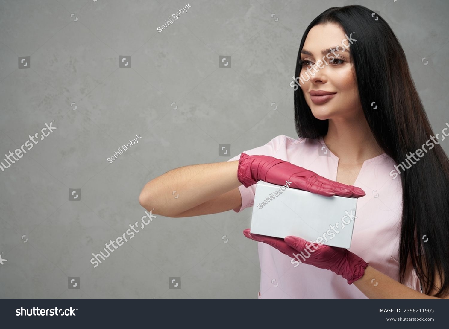 Happy female cosmetologist holding white box with both hands. Portrait of dark haired pretty woman wearing pink gloves and uniform, showing small box against grey wall. Beauty, cosmetology concept. #2398211905