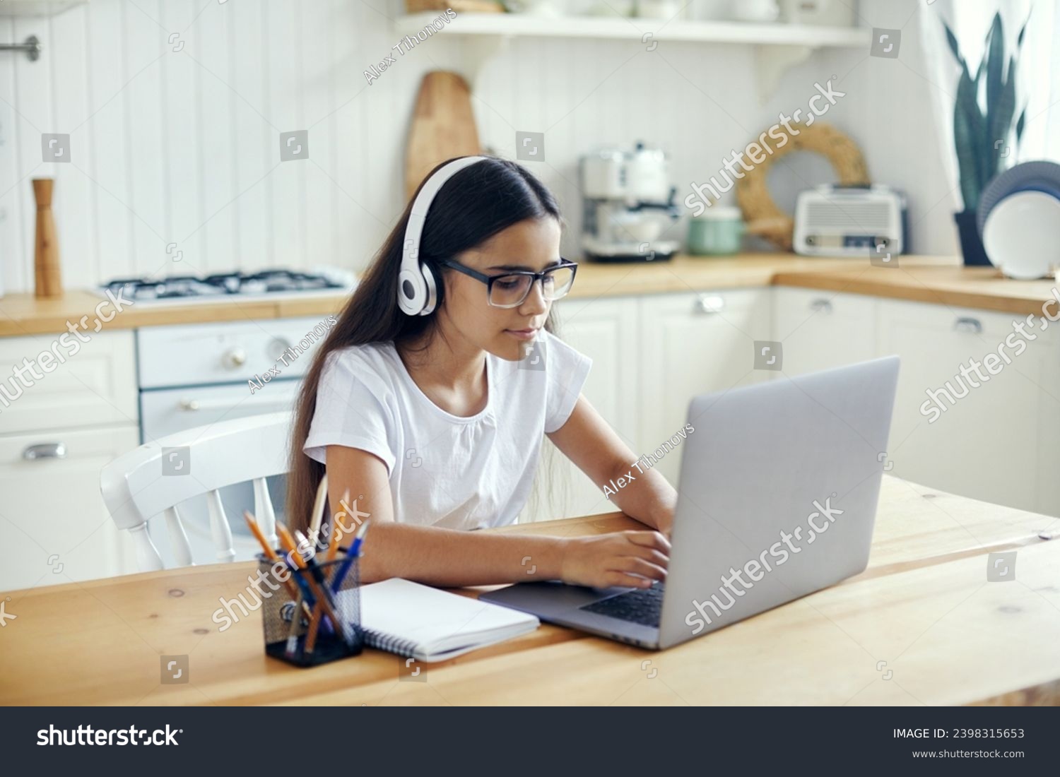 Pre-teen 12s girl in headphones and eyeglasses sit at table e-learns, listen online course, audio lesson, get new knowledge, skills using internet and modern tech. Development, education, eye-sight #2398315653