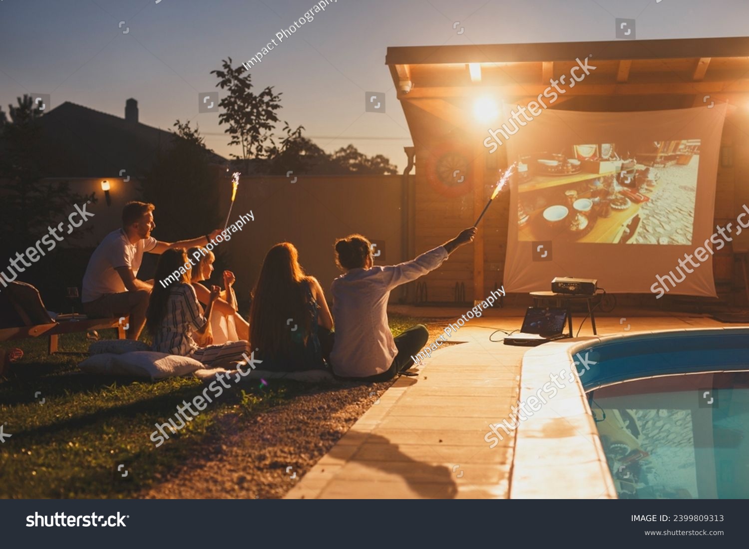 Group of friends having a surprise birthday party in an open air cinema, watching a movie by the swimming pool and waving with sparklers #2399809313