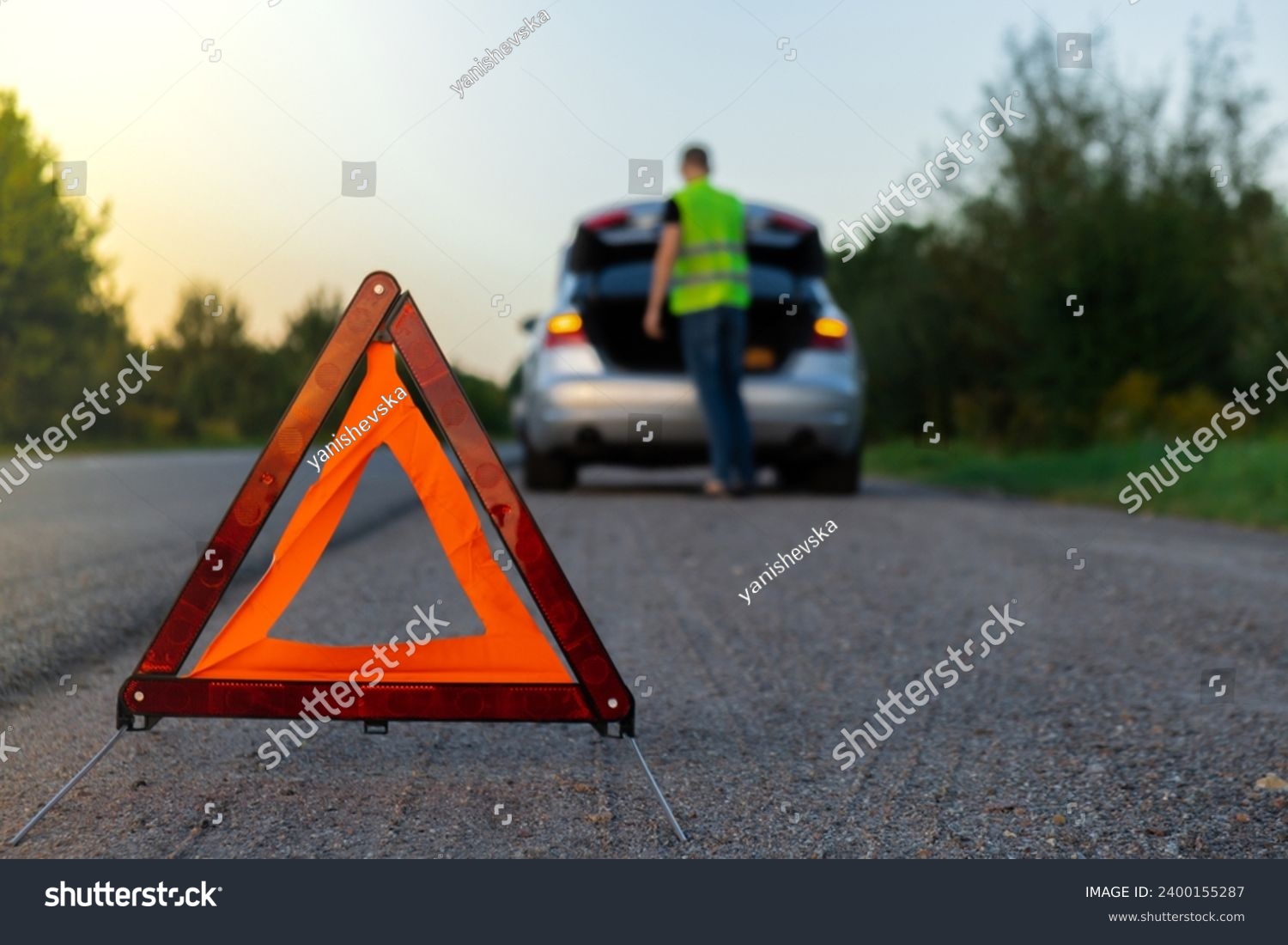 Broken silver luxury car emergency accident. Man driver installing red triangle stop sign on road. Sport automobile turned on blinkers technical problems on the road. Safety procedure vehicle broken #2400155287