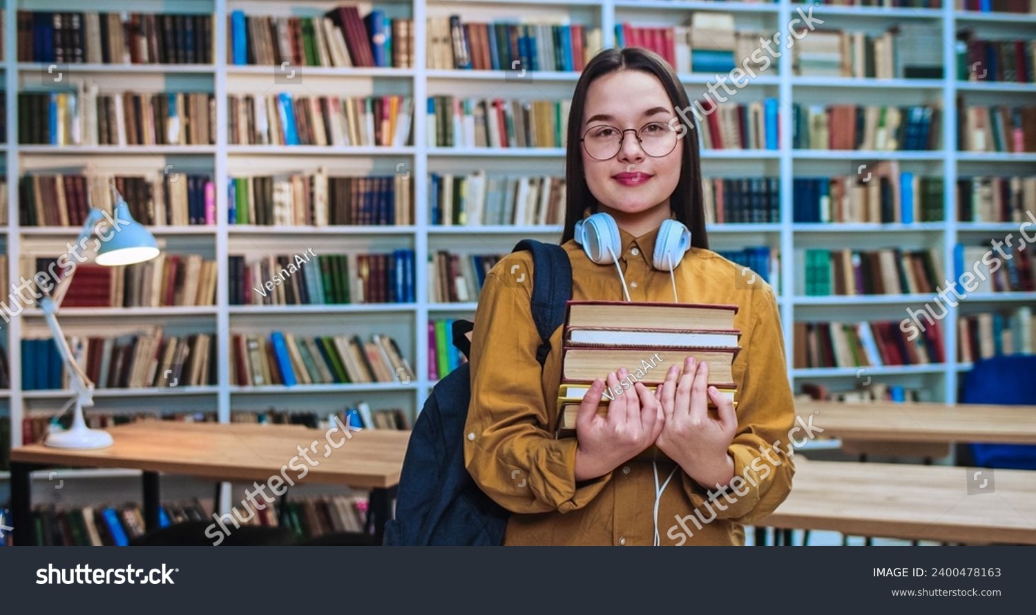 Portrait of cute brunette student with white headsets, black backpack and lots of book in hands smiling and looking in camera in library. #2400478163