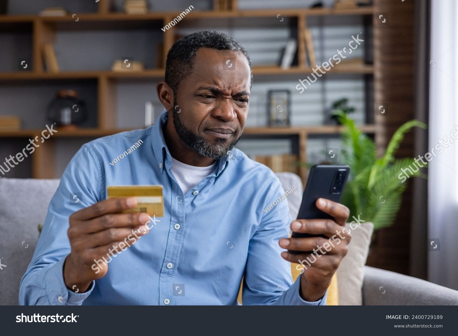 Upset and sad man sitting at home on couch in living room, cheated senior mature african american man displeased holding phone and bank credit card, rejected fund transfer error, account block. #2400729189