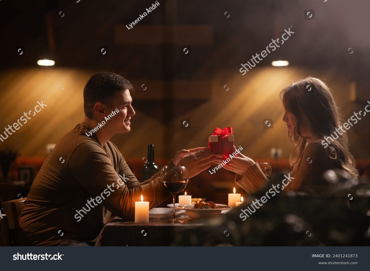 Happy young couple celebrating Valentines day having romantic dinner at home. Loving man giving gift box his woman. Copy space #2401241873