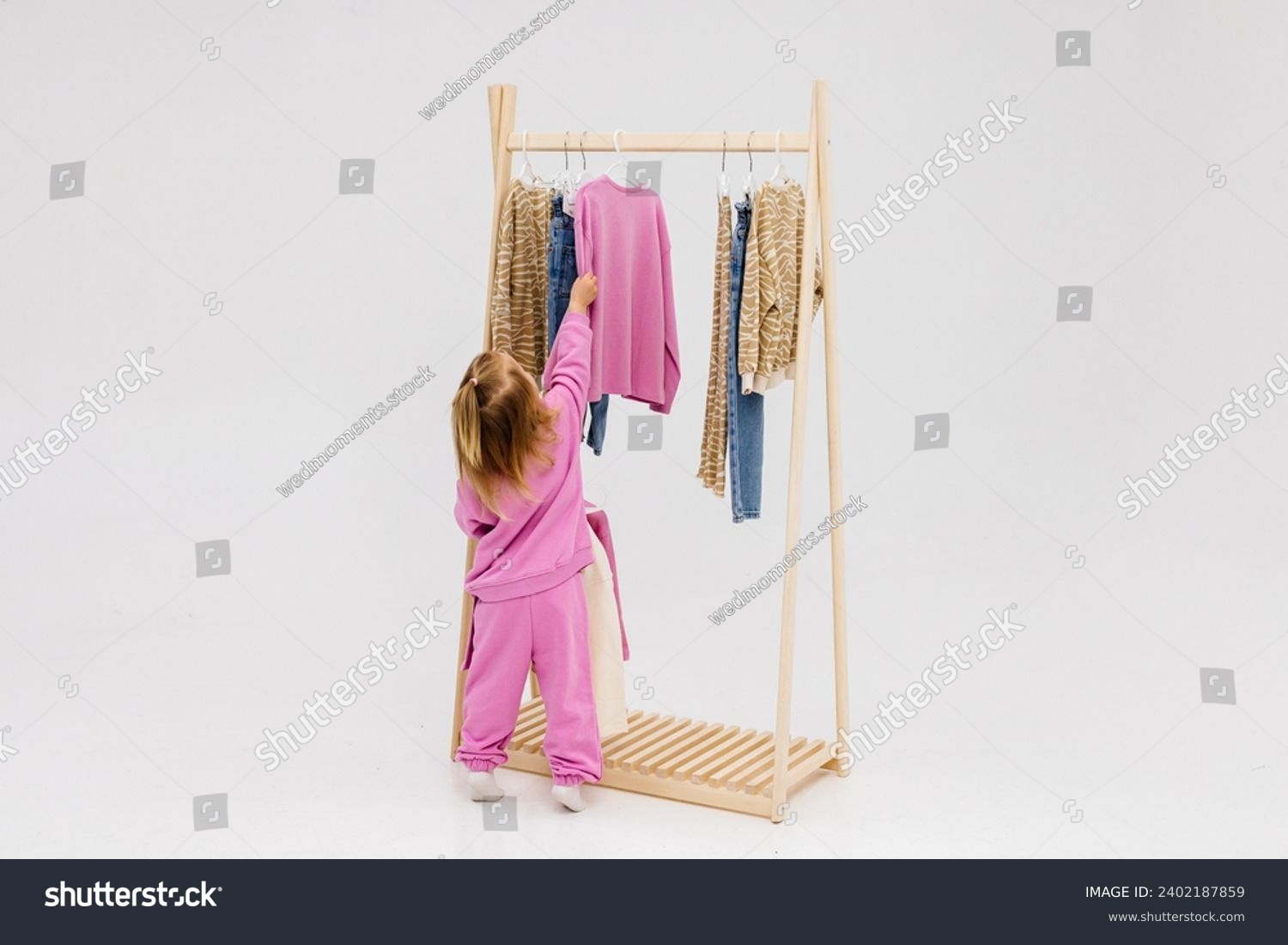 A child, a little girl, stands near the closet, chooses clothes against a light background. Dressing room with clothes on hangers. Wardrobe of children's and stylish clothes. Montessori wardrobe. #2402187859