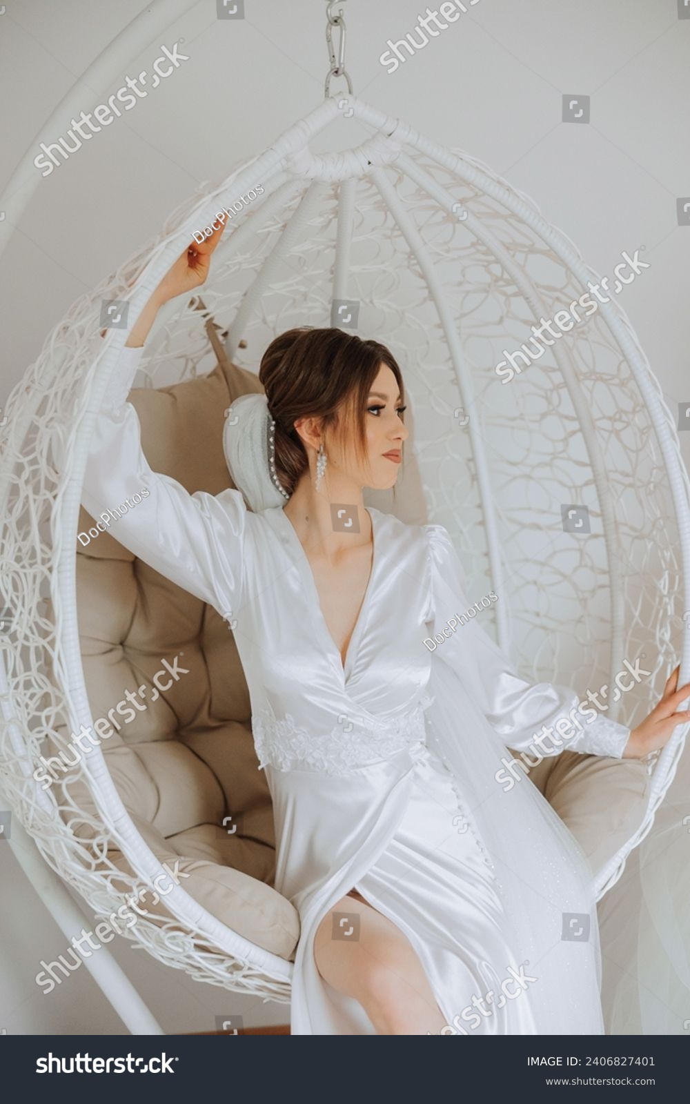 Morning of the bride in boudoir style. A woman bride in a white robe sits on a wicker swing near her wedding dress on a mannequin. Wedding day. #2406827401