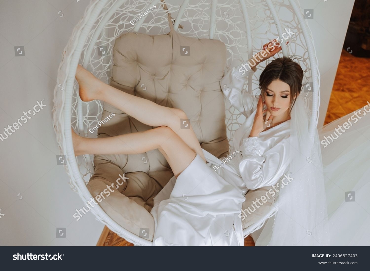Morning of the bride in boudoir style. A woman bride in a white robe sits on a wicker swing near her wedding dress on a mannequin. Wedding day. #2406827403