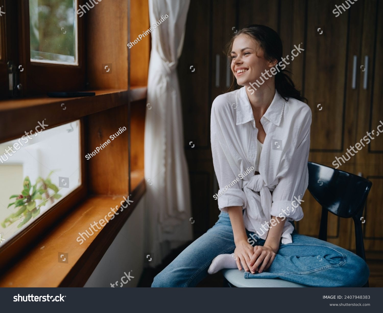 Woman sitting at home by a wooden window with a smile on a chair in homemade comfortable clothes and looking at the landscape, spring mood, women's day, rest on the weekend. #2407948383