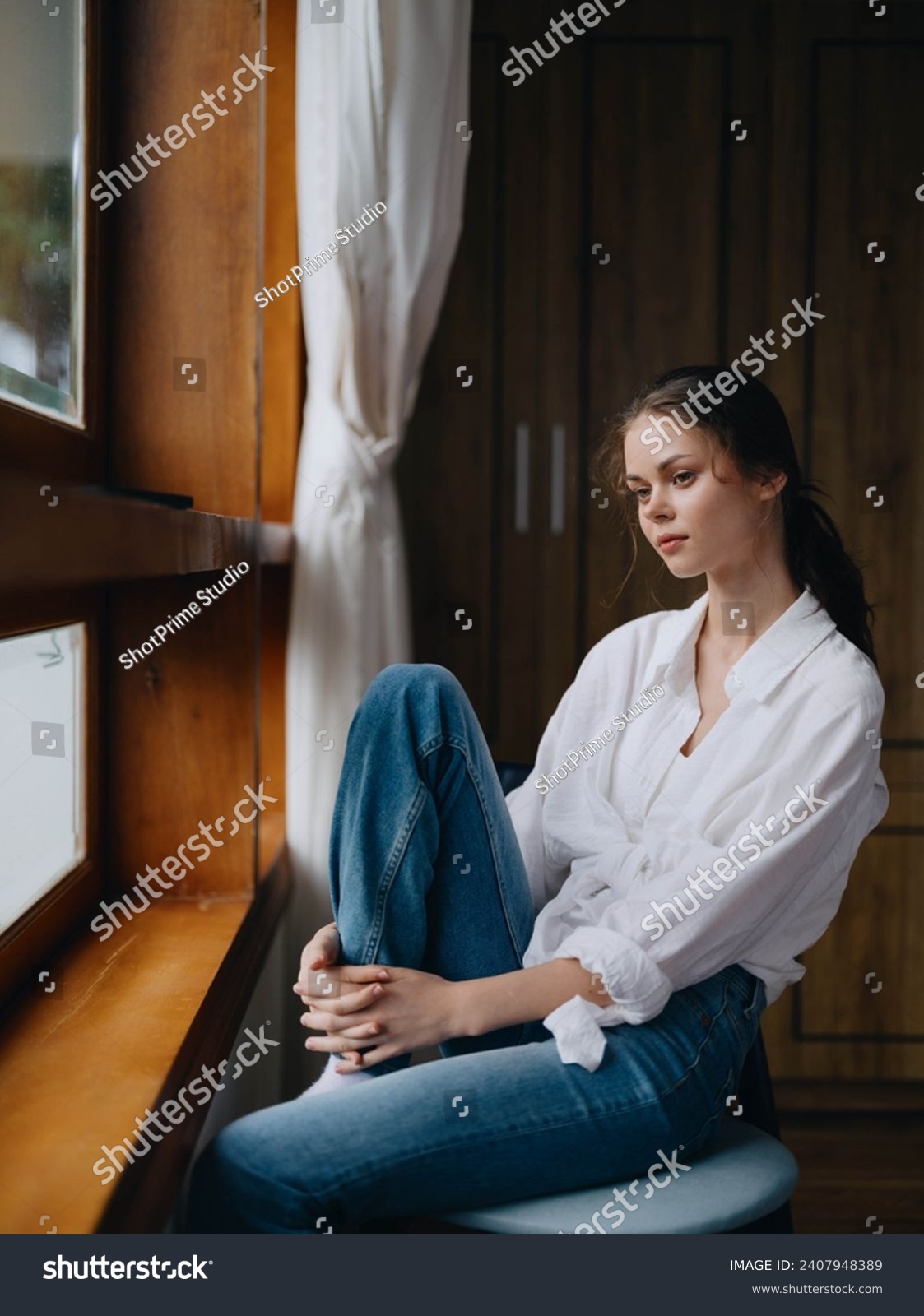 Woman sitting at home by a wooden window with a smile on a chair in homemade comfortable clothes and looking at the landscape, spring mood, women's day, rest on the weekend. #2407948389