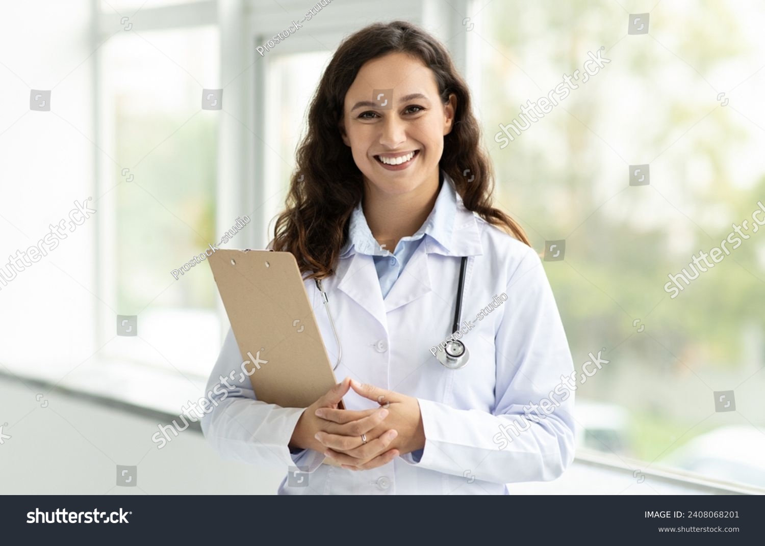 Cheerful millennial lady doctor in white coat, with stethoscope and clipboard standing next to window at clinic, hospital, copy space. Medical exam, health care consultation, prescription #2408068201
