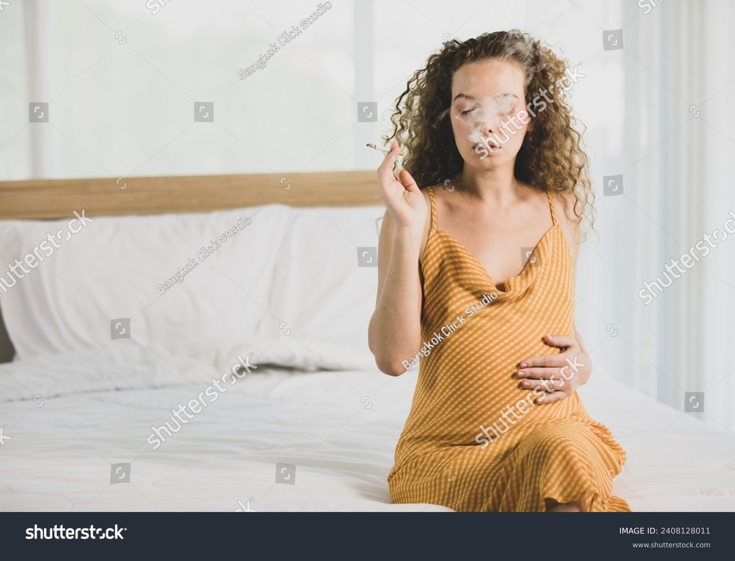 Curly hairstyle young unhappy unhealthy bad behavior Caucasian pregnancy mother in maternity long dress cloth sit on bed in bedroom holding smoking cigarette taking risk and danger to unborn child. #2408128011