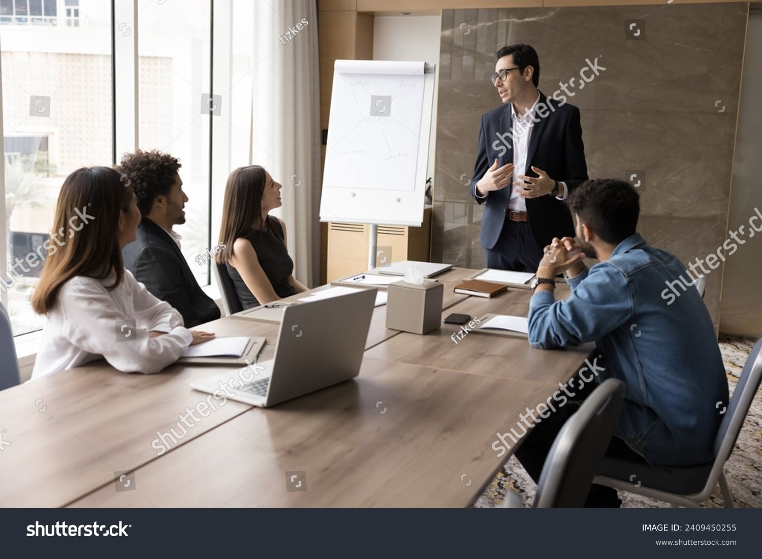 Serious young business leader man speaking to diverse team of colleagues, standing at meeting table, telling project plan, management strategy, ideas for brainstorming #2409450255