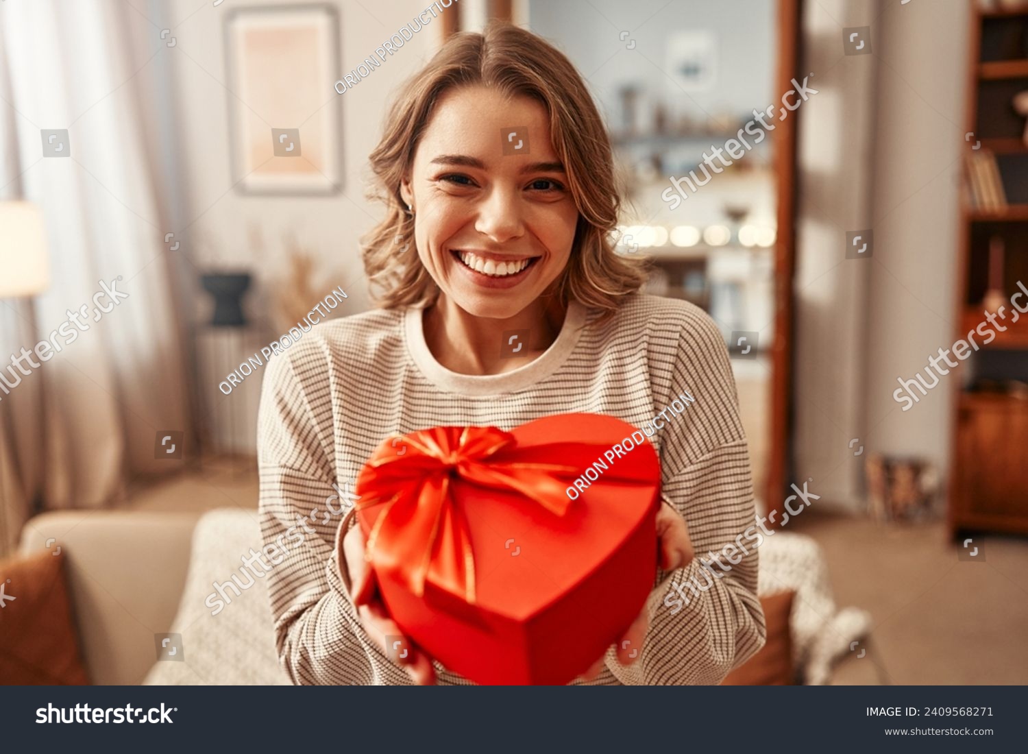 Happy Valentine's Day. Happy smiling woman holding red heart shaped gift box with bow in living room at home. #2409568271