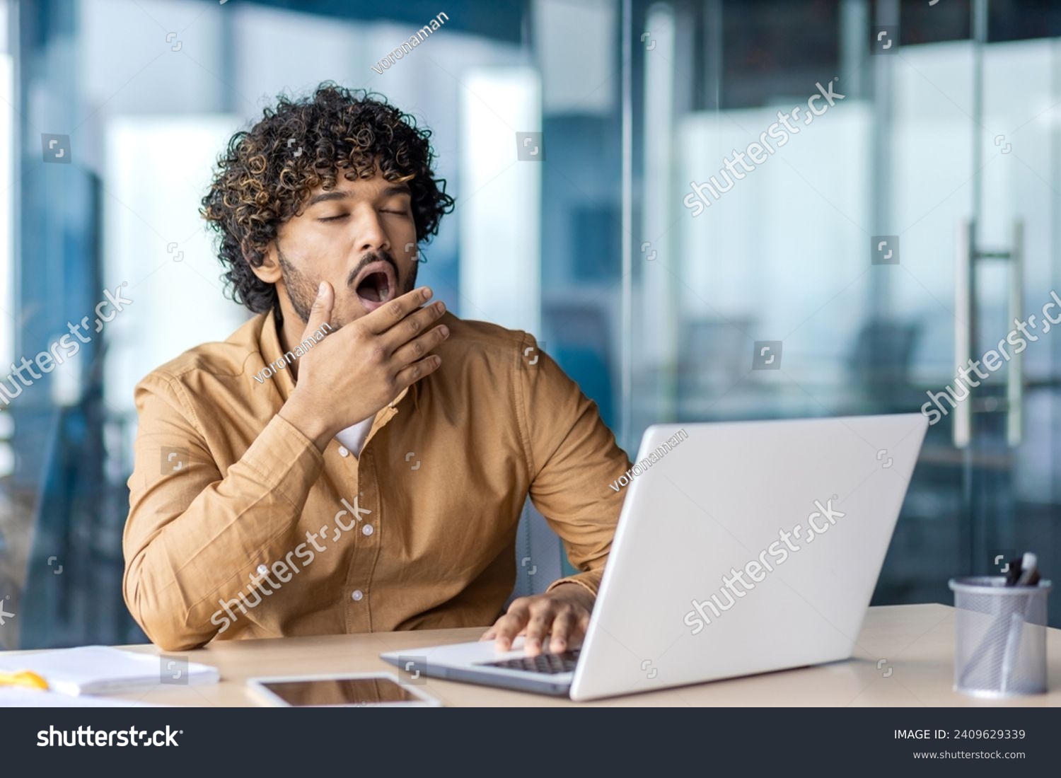 Young overworked and overtired businessman yawning, working inside office with laptop, boring long routine work, man in casual clothes. #2409629339