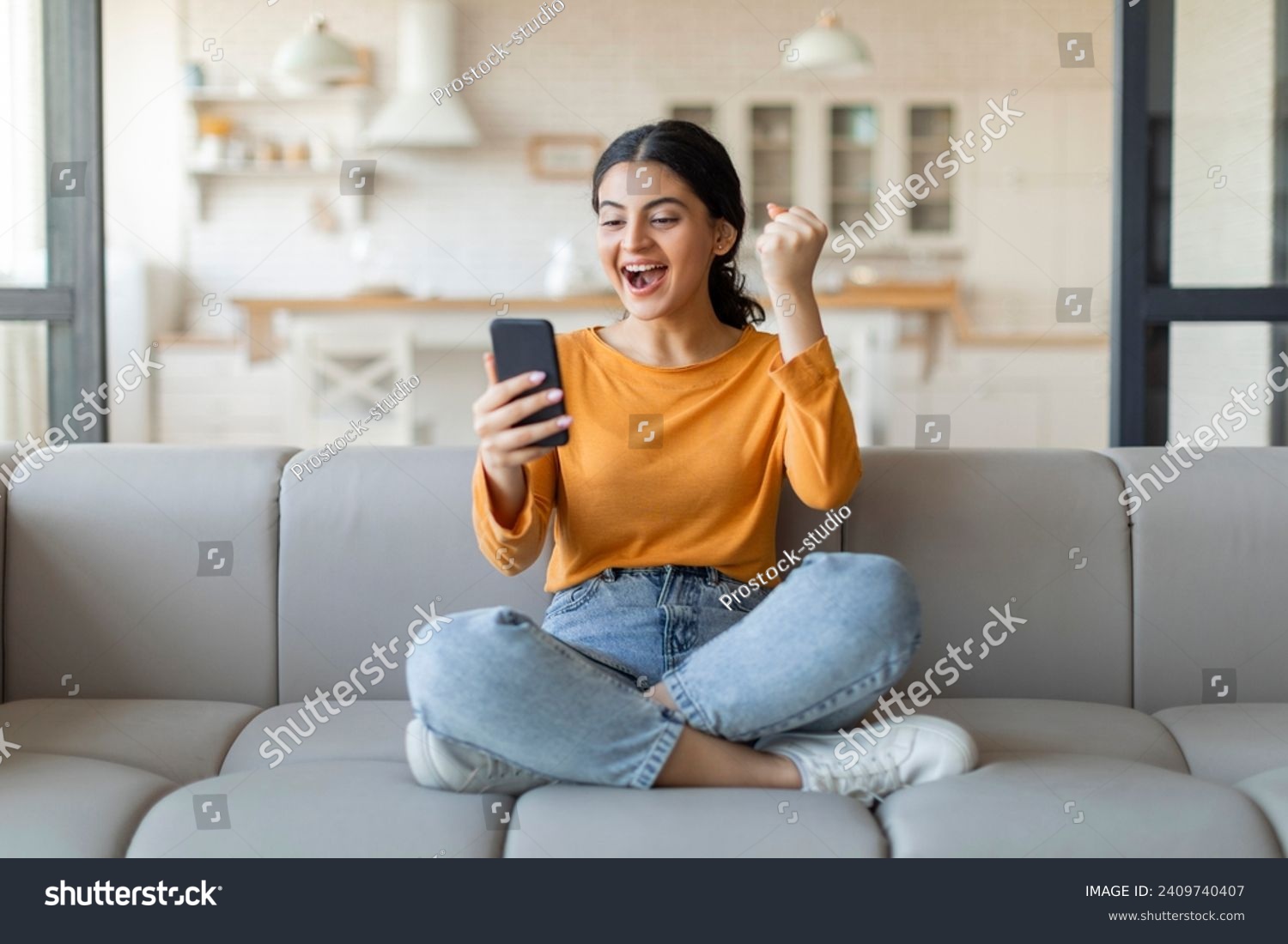 Joyful young indian woman with smartphone celebrating victory. happy excited female sitting on couch at home, holding mobile phone and shaking fist, cheering success and online win, got good news #2409740407