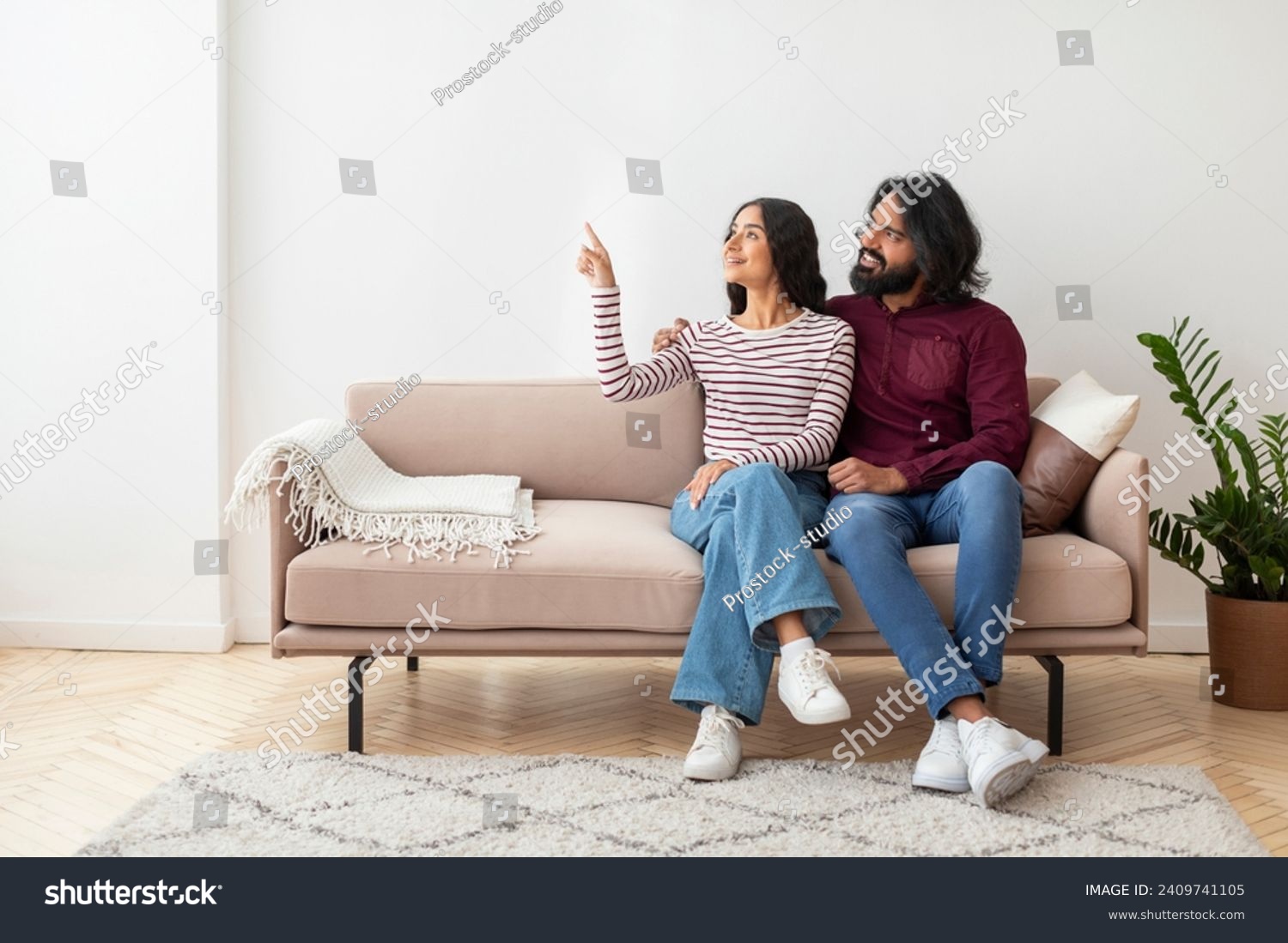 Lovely smiling young indian couple wearing casual outfit sitting on couch at home, brunette woman pointing at copy space, showing her husband nice offer, living room interior. Mortgage, moving #2409741105