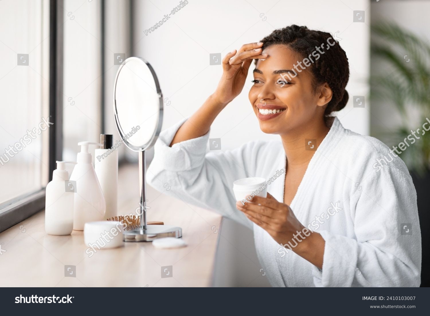 Beautiful Black Woman Sitting Near Mirror And Applying Moisturising Cream On Face, Attractive African American Lady Wearing Bathrobe Nourishing Skin After Bath, Making Beauty Routine At Home #2410103007
