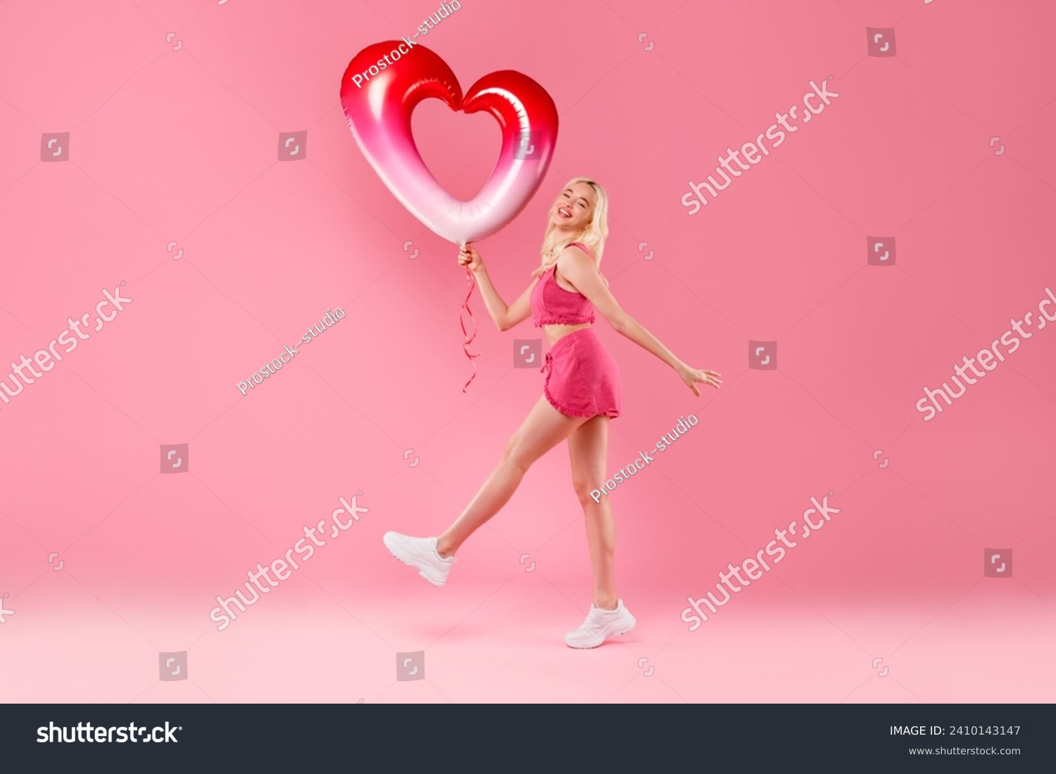 Blonde girl joyfully dancing, holding a red and white heart balloon, in pink fuzzy outfit with sneakers on a pink background, feeling elated, full length #2410143147