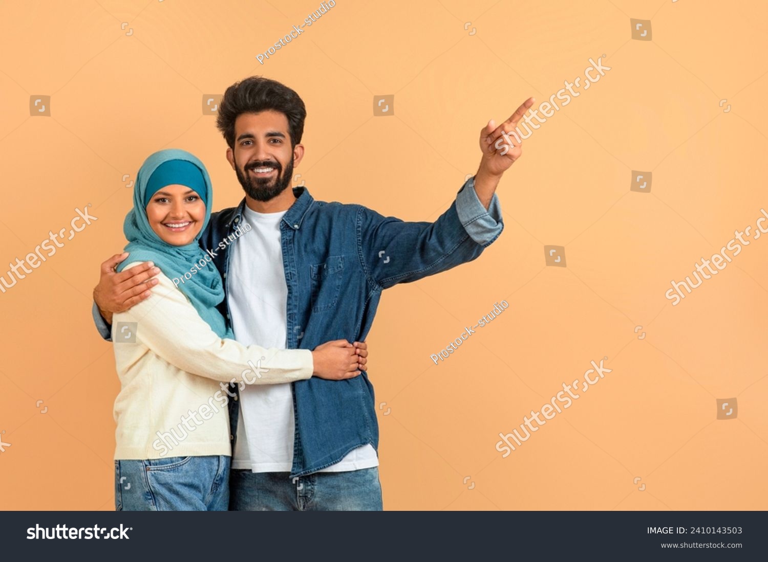 Arab muslim couple embracing and pointing aside at free space, smiling young islamic spouses sharing moment of discovery, demonstrating place for your ad, standing over soft beige studio background #2410143503