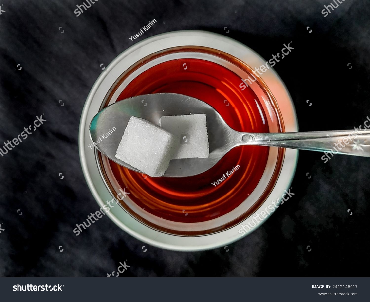 Sugar cubes on a teaspoon with a cup of tea on a black cloth background #2412146917