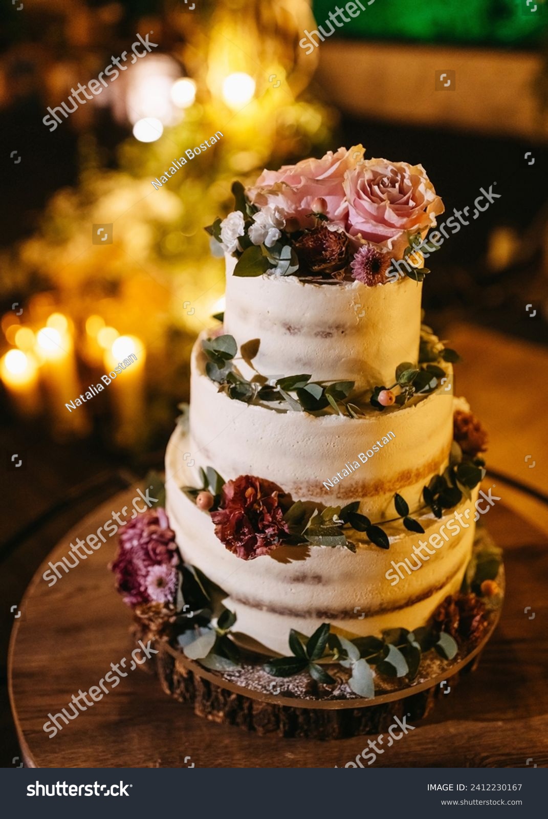 A three-tiered wedding cake adorned with fresh flowers and greenery, set against a backdrop of warm candlelight. #2412230167