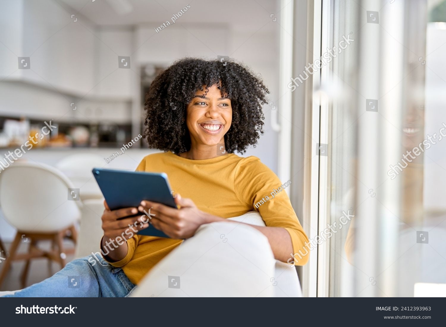 Happy African American lady using tab device looking away on sofa at home. Smiling pretty young woman sitting on couch relaxing looking away at window holding digital table in kitchen. #2412393963