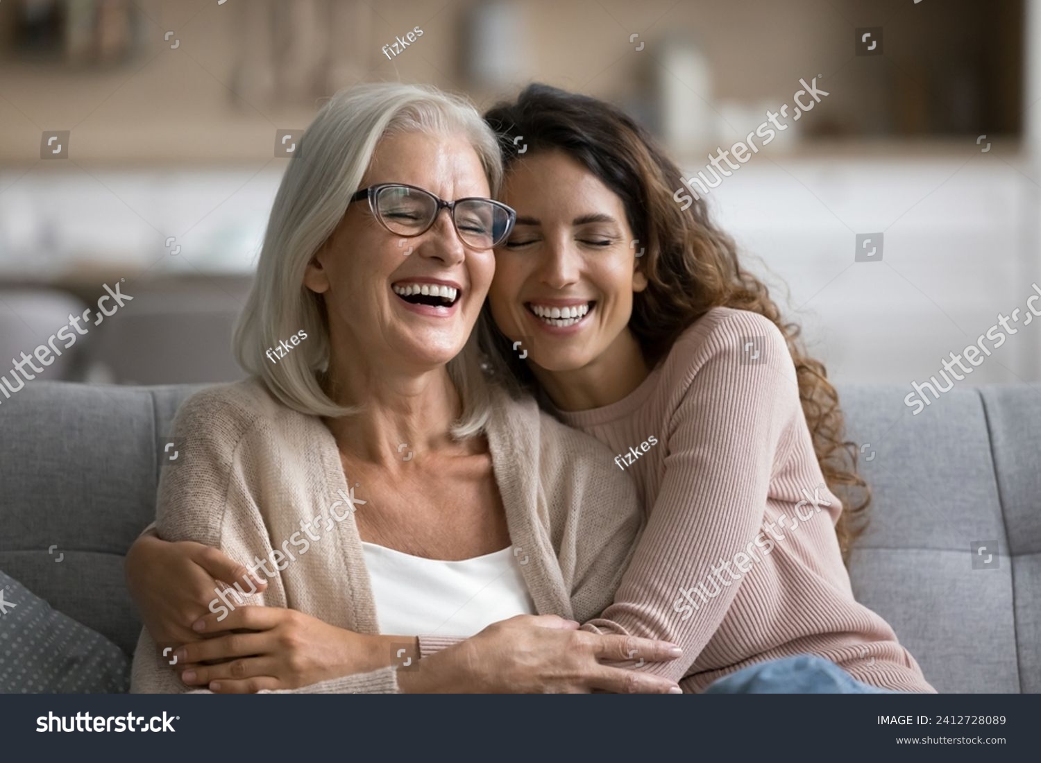 Happy excited senior mom and adult daughter woman having fun at home, hugging with heads touch, sitting on sofa, laughing with closed eyes, enjoying funny talk, leisure, family affection #2412728089