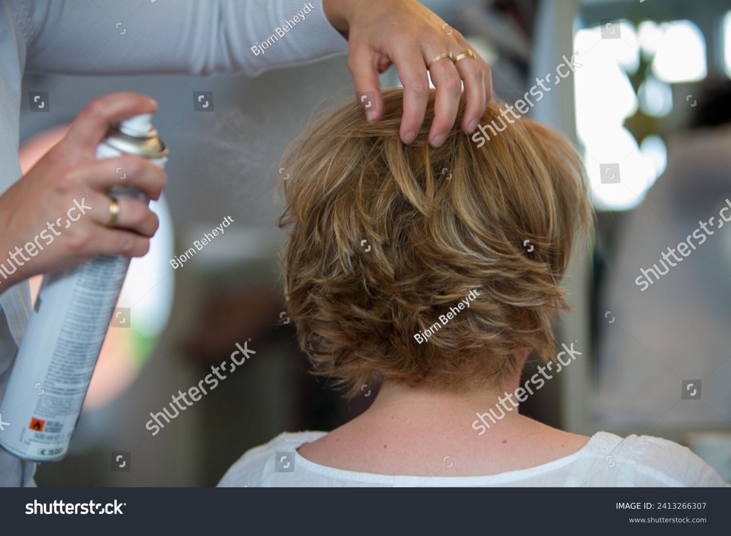 The image captures a moment of hairstyling where hands gently sculpt a short, layered haircut while applying hairspray to set the style in place. The hairstylist's hands are adorned with a wedding #2413266307