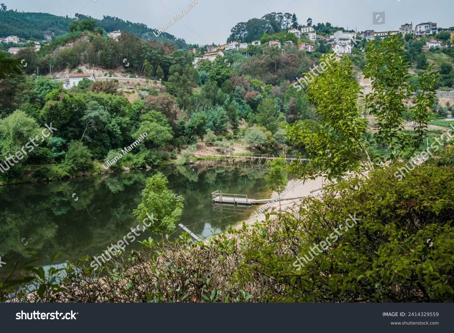 Green leafy bushes framing the Mondego river with wooden bridge in the valley with houses on the hill, Penacova PORTUGAL #2414329559