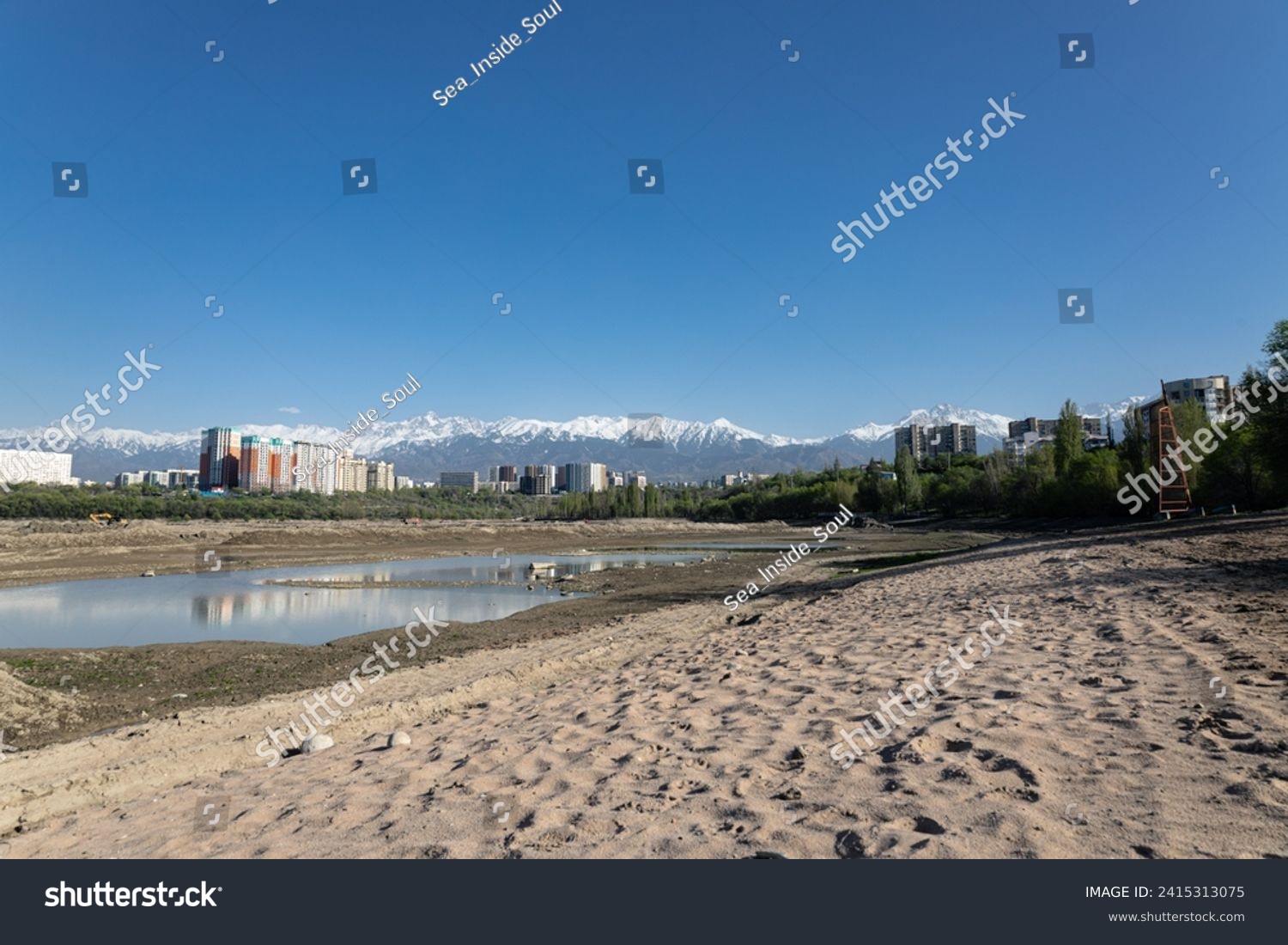 Storage reservoir Lake Sayran, Almaty, Kazakhstan. Empty City sand beach with drained pond. Residential apartment buildings and high snow-capped mountains in background. Recreation place for citizens #2415313075