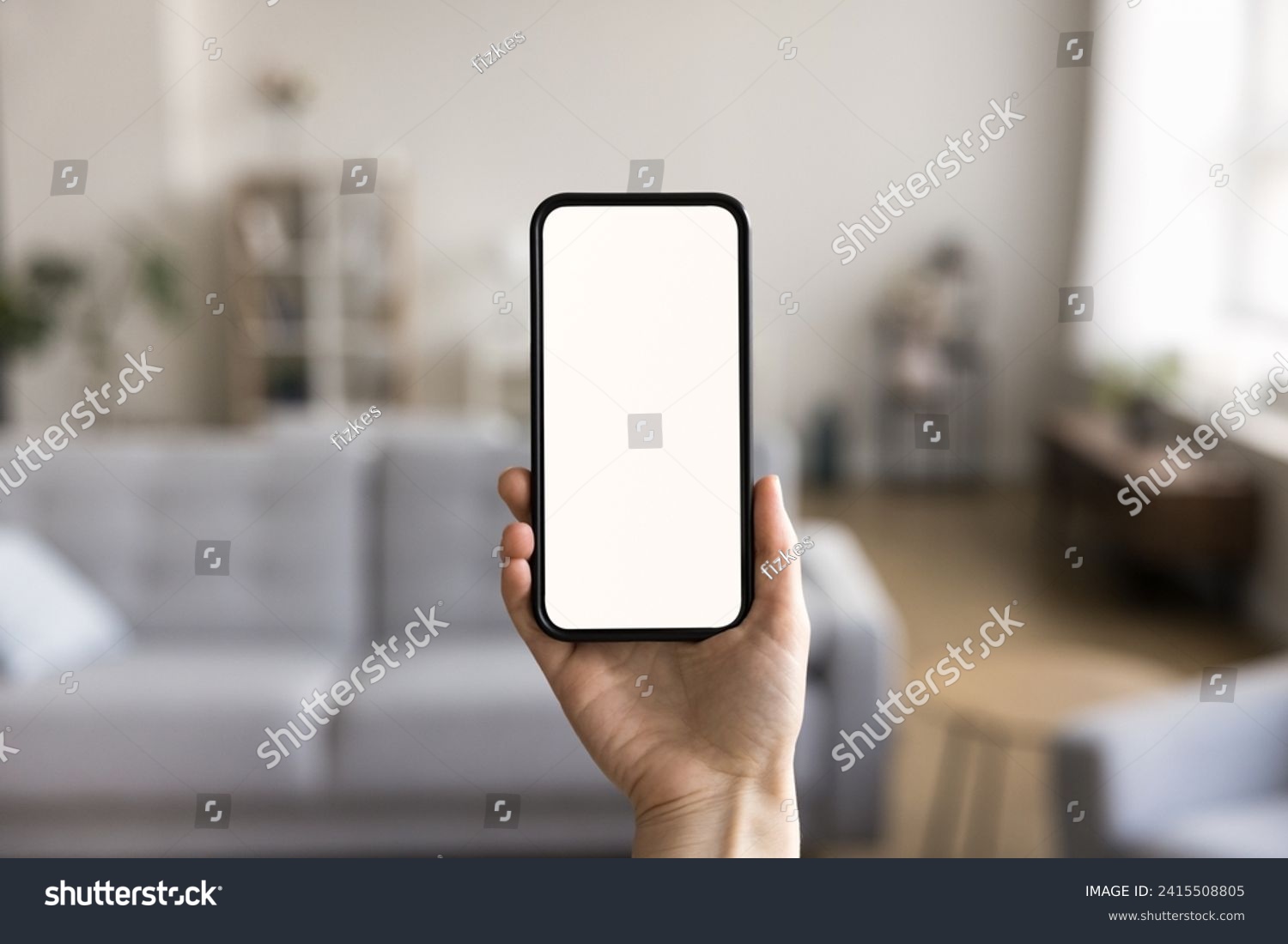 Close up shot of unknown female hand holding modern smartphone with white mock up blank screen, living room on background. Advertisement of new mobile application ad, smart home app, comfort living #2415508805