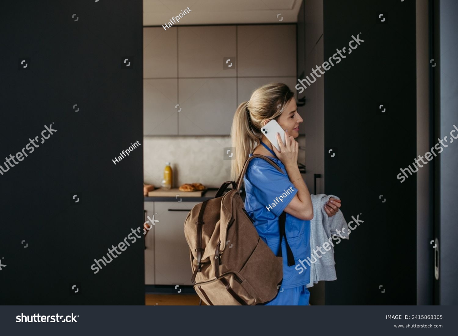 Female doctor getting ready for work, reading message on smartphone, leaving house in scrubs with backpack. Work-life balance for healthcare worker. #2415868305