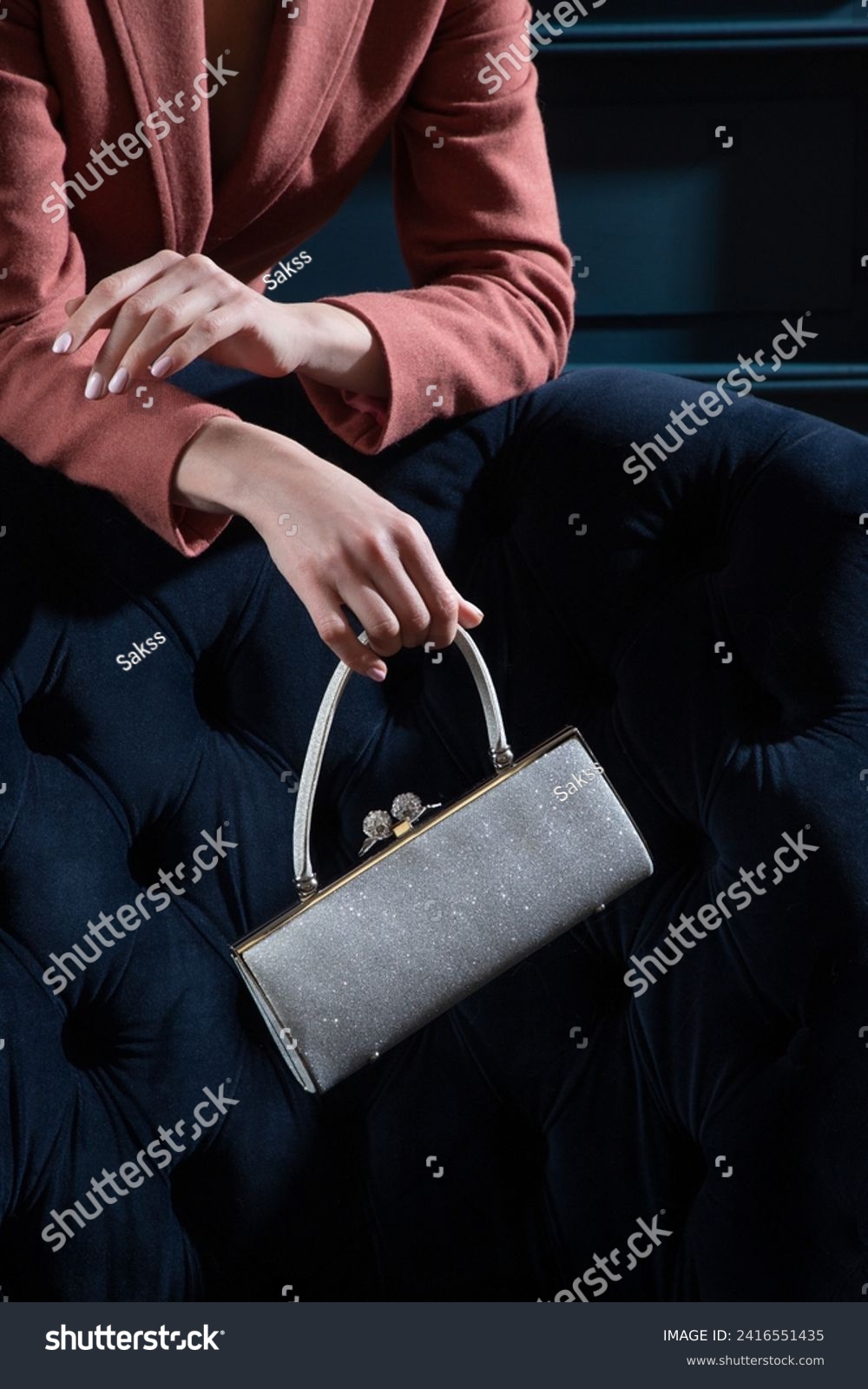Silver metallic sparkling purse held by woman hands on a blue velour armchair background. Creative shiny woman hand bag studio photography. #2416551435