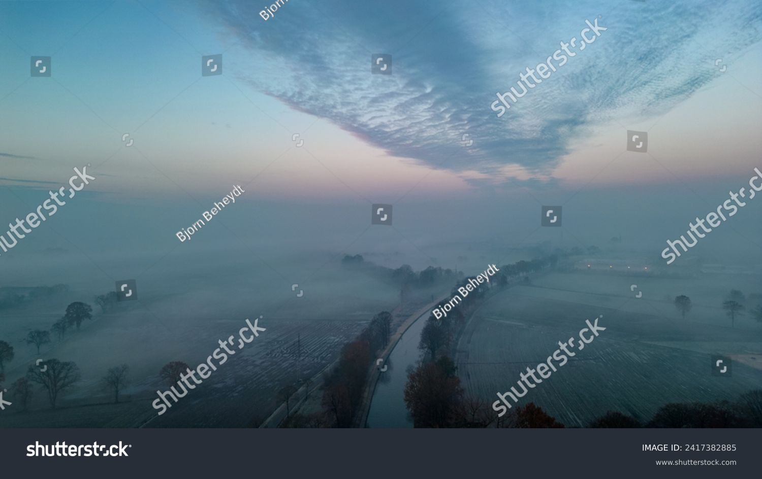 This evocative aerial image captures the tranquil beauty of dawn as it breaks through a layer of mist over a frosty winter landscape. The sky, filled with a dramatic formation of clouds, gradually #2417382885