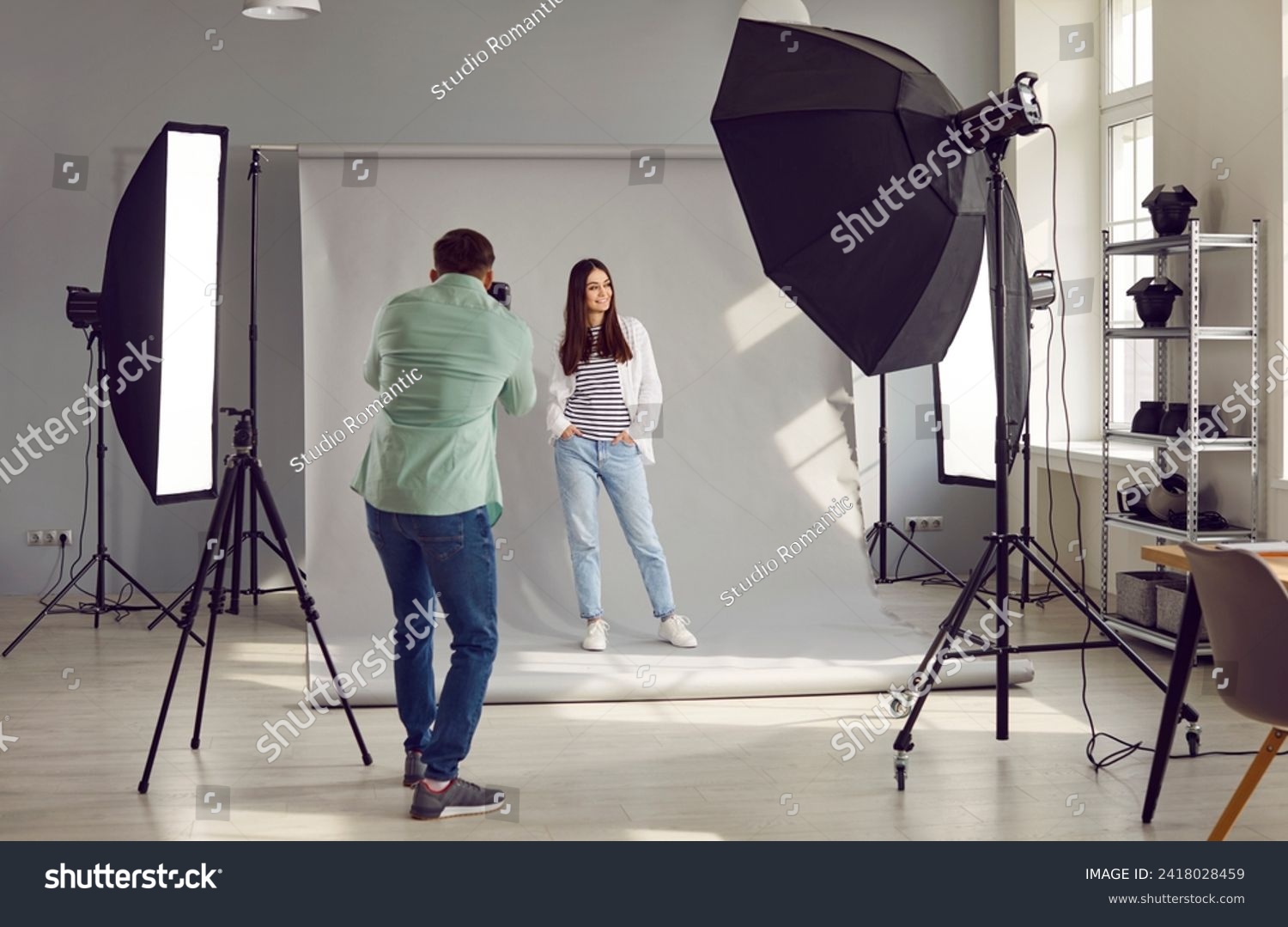 Beautiful model posing for male photographer in studio. Rare view of photographer taking pictures of female model with digital camera in photo studio with professional photographic equipment #2418028459