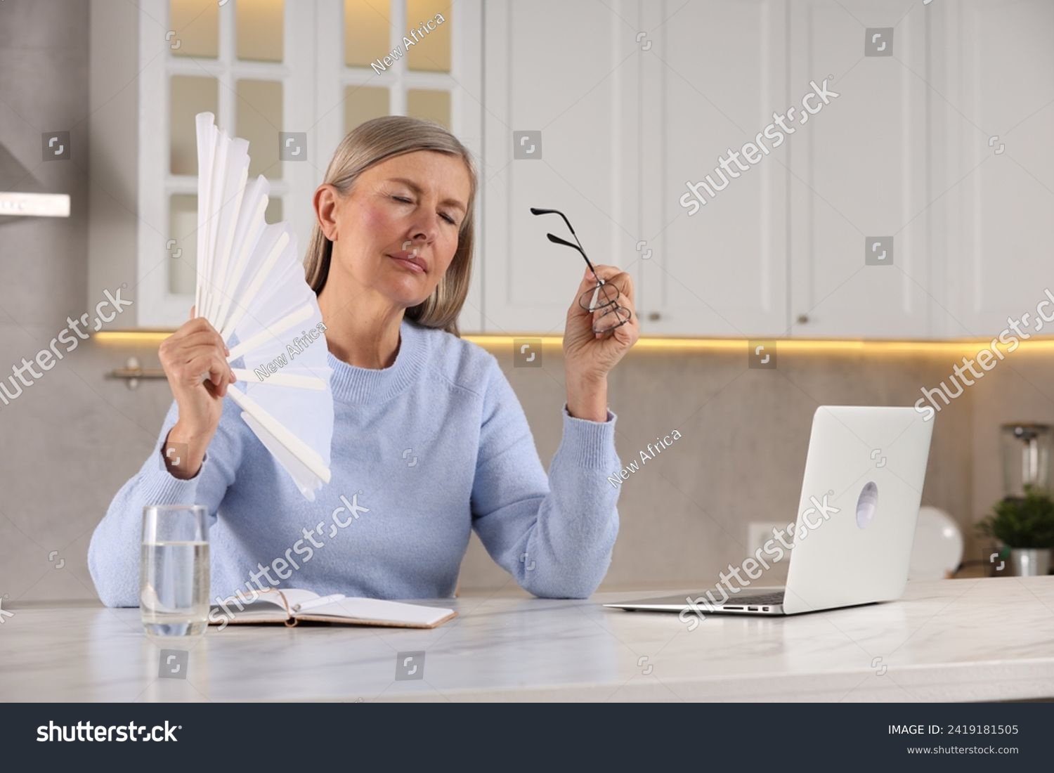 Menopause. Woman waving hand fan to cool herself during hot flash at table in kitchen #2419181505