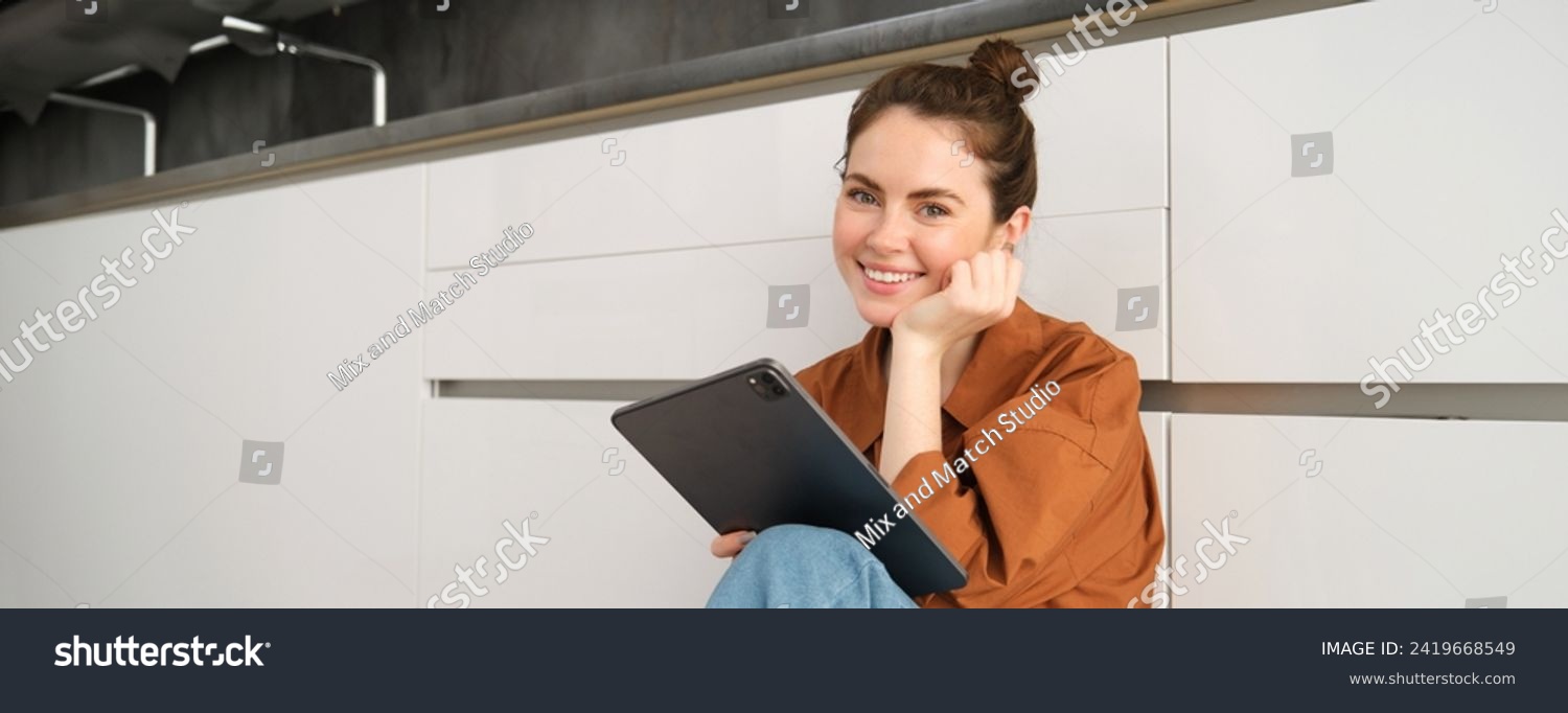 Portrait of young beautiful woman sitting on kitchen floor with digital tablet, browsing news feed, social media app on gadget, smiling and looking happy. #2419668549
