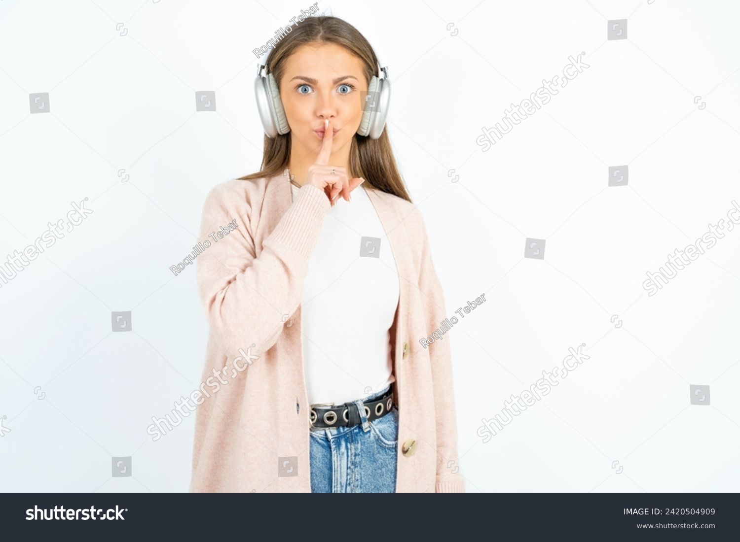 Young beautiful woman standing over white studio background making hush gesture with finger on her lips wearing  wireless headphones. Be quiet. #2420504909